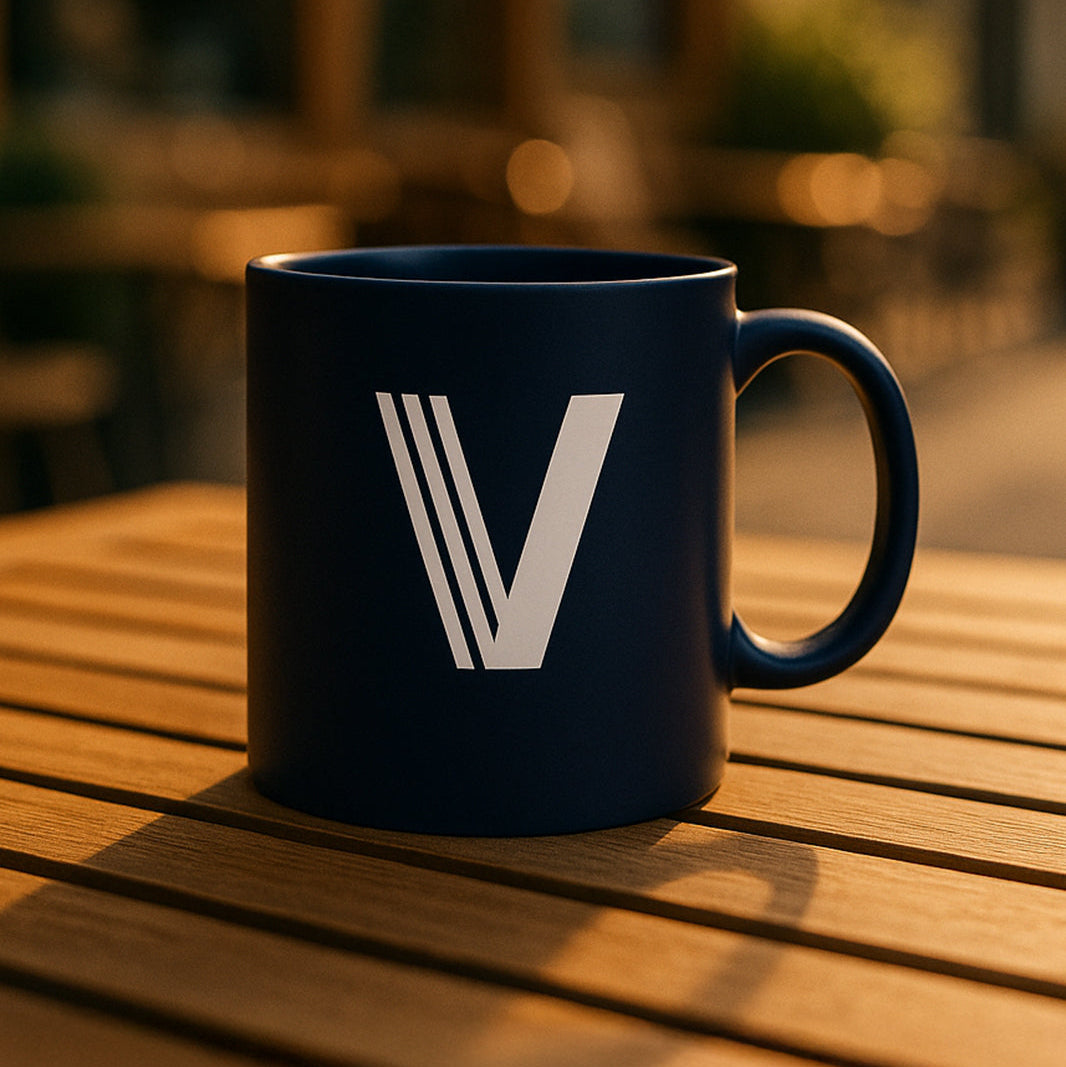 Dark blue mug with a white V logo, sitting on a wooden slatted table in warm sunlight.