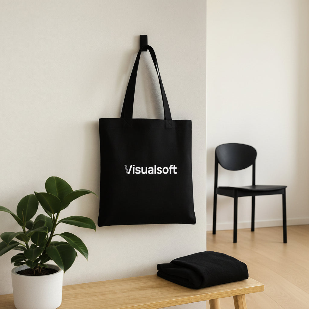 Black tote bag with Visualsoft logo hanging on a white wall, next to a plant and a wooden bench.